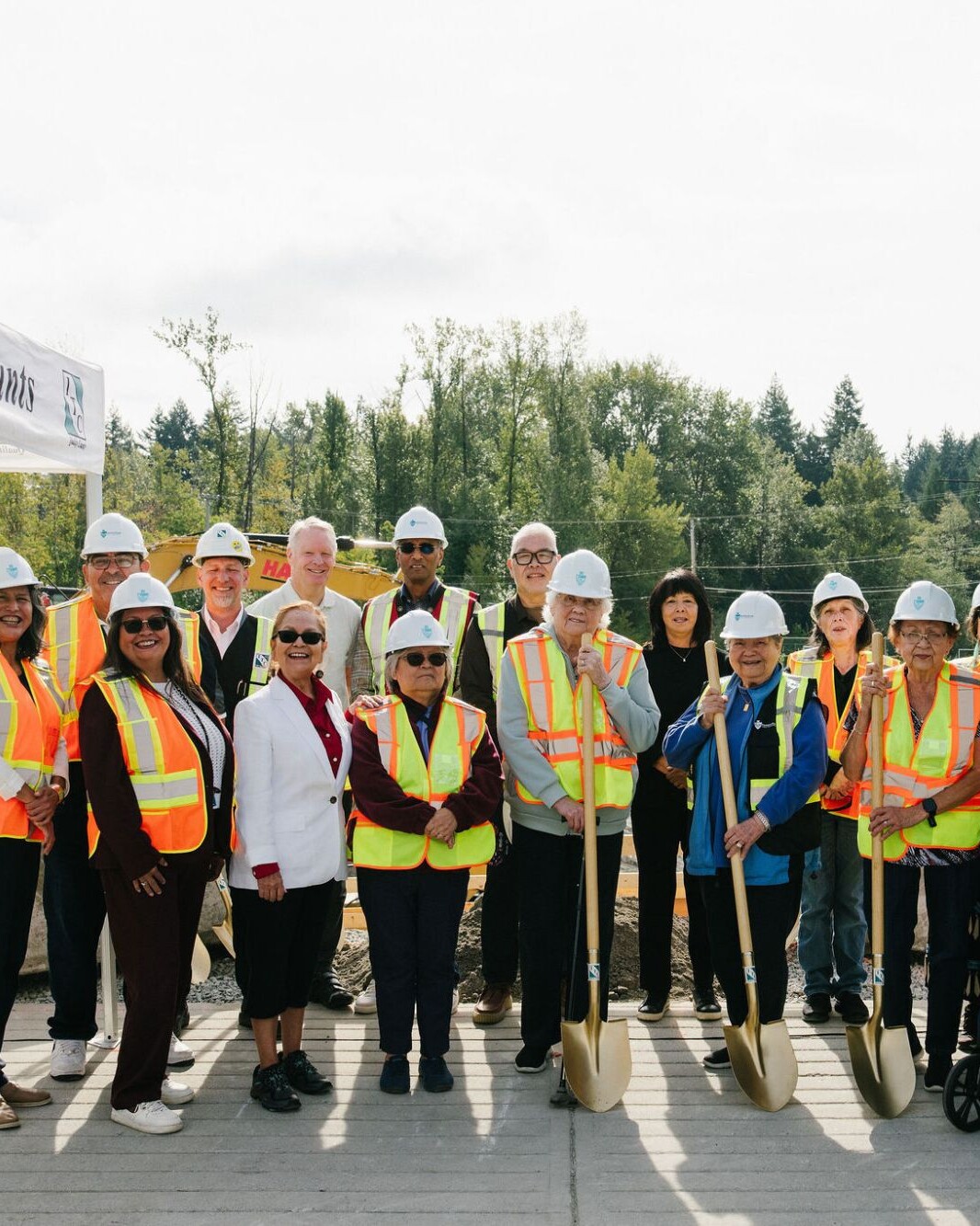 Musqueam Elders, leaders and guests in hard hats and safety vests, holding ceremonial gold shovels at the East Wind groundbreaking at leləm̓ village.