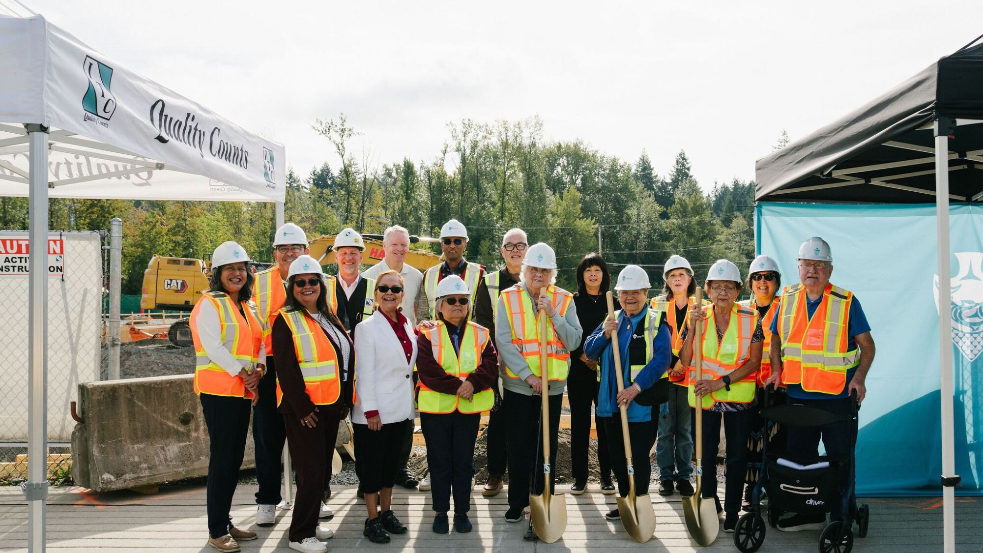Musqueam Elders, leaders and guests in hard hats and safety vests, holding ceremonial gold shovels at the East Wind groundbreaking at leləm̓ village.