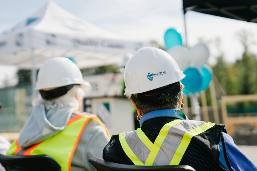 Two guests in hard hats and safety vests, one hard hat marked Musqueam Capital Corporation, watching the East Wind groundbreaking ceremony.