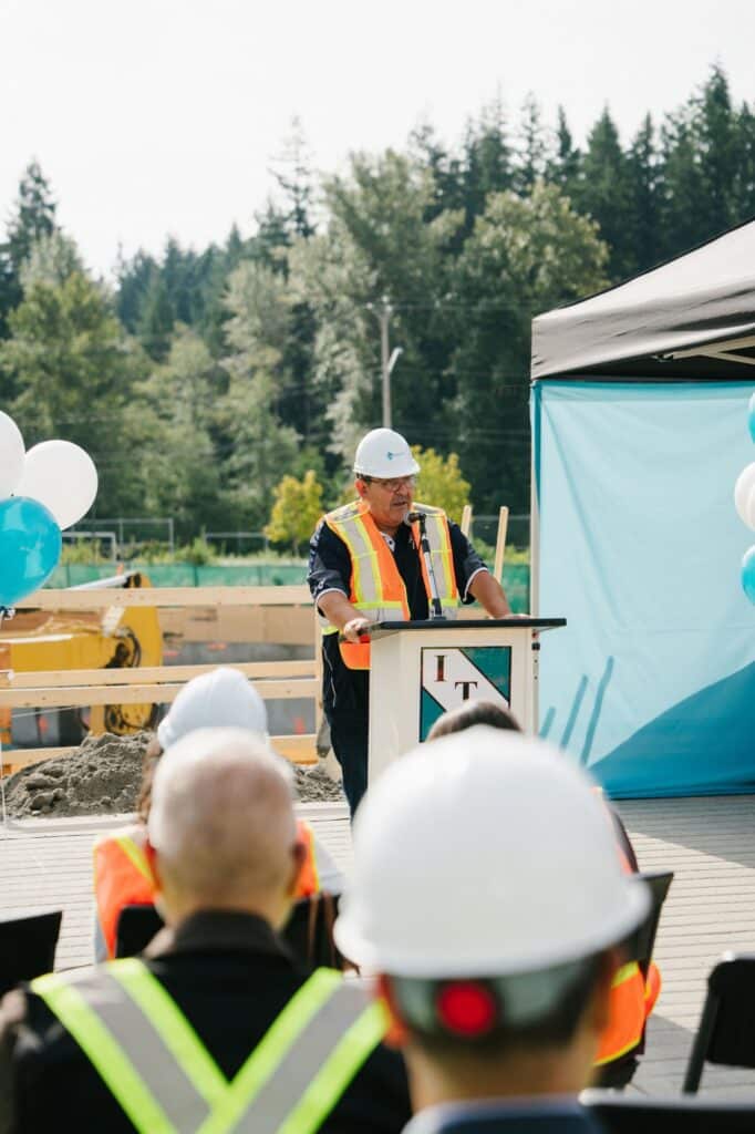 A speaker in a hard hat and safety vest addresses guests from a podium at the East Wind groundbreaking ceremony.